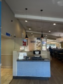 A restaurant reception area with a check-in counter featuring a blue brick-patterned front. The counter has a cash register and some signs, one indicating 'Pick up here.' Above the counter hang two black pendant lights. The restaurant interior includes tables and booths visible to the right.