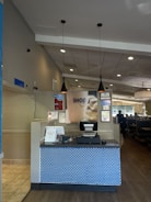 A restaurant reception area with a check-in counter featuring a blue brick-patterned front. The counter has a cash register and some signs, one indicating 'Pick up here.' Above the counter hang two black pendant lights. The restaurant interior includes tables and booths visible to the right.