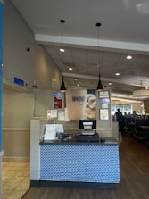 A restaurant reception area with a check-in counter featuring a blue brick-patterned front. The counter has a cash register and some signs, one indicating 'Pick up here.' Above the counter hang two black pendant lights. The restaurant interior includes tables and booths visible to the right.