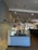 A restaurant reception area with a check-in counter featuring a blue brick-patterned front. The counter has a cash register and some signs, one indicating 'Pick up here.' Above the counter hang two black pendant lights. The restaurant interior includes tables and booths visible to the right.