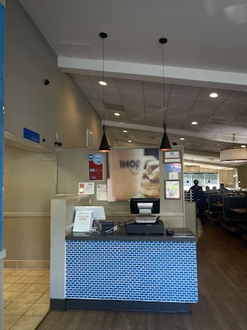 A restaurant reception area with a check-in counter featuring a blue brick-patterned front. The counter has a cash register and some signs, one indicating 'Pick up here.' Above the counter hang two black pendant lights. The restaurant interior includes tables and booths visible to the right.