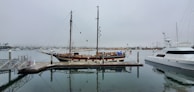 A calm marina scene featuring a wooden sailing ship docked at a pier. The ship has a tall mast with rigging and is accompanied by a modern yacht on the right side. The sky is overcast, reflecting a grey tone on the water, which is still and mirrors the boats.