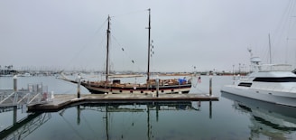 A calm marina scene featuring a wooden sailing ship docked at a pier. The ship has a tall mast with rigging and is accompanied by a modern yacht on the right side. The sky is overcast, reflecting a grey tone on the water, which is still and mirrors the boats.
