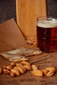 A glass of dark beer sits on a wooden surface next to dried fish and a pile of nuts, all arranged on top of printed paper. A wooden cutting board is visible in the background.