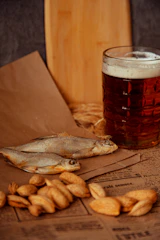 Close-up of assorted dried fish snacks arranged on rustic wooden board with hops and beer glasses in the background