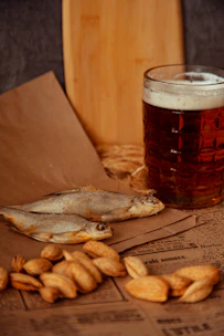 Close-up of assorted dried fish snacks arranged on rustic wooden board with hops and beer glasses in the background