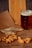 A glass of dark beer sits on a wooden surface next to dried fish and a pile of nuts, all arranged on top of printed paper. A wooden cutting board is visible in the background.
