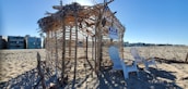 A beach scene with a makeshift hut made of wooden sticks and dried palm leaves. Two white Adirondack chairs are placed in front of the structure, which has a 'For Rent' sign hanging. Surrounding the hut is an expanse of sandy beach, and modern beachfront homes are visible in the background under a clear blue sky.