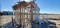 A beach scene with a makeshift hut made of wooden sticks and dried palm leaves. Two white Adirondack chairs are placed in front of the structure, which has a 'For Rent' sign hanging. Surrounding the hut is an expanse of sandy beach, and modern beachfront homes are visible in the background under a clear blue sky.