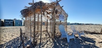 A beach scene with a makeshift hut made of wooden sticks and dried palm leaves. Two white Adirondack chairs are placed in front of the structure, which has a 'For Rent' sign hanging. Surrounding the hut is an expanse of sandy beach, and modern beachfront homes are visible in the background under a clear blue sky.
