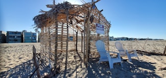 A beach scene with a makeshift hut made of wooden sticks and dried palm leaves. Two white Adirondack chairs are placed in front of the structure, which has a 'For Rent' sign hanging. Surrounding the hut is an expanse of sandy beach, and modern beachfront homes are visible in the background under a clear blue sky.