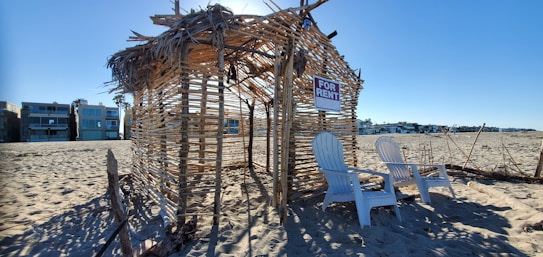 A beach scene with a makeshift hut made of wooden sticks and dried palm leaves. Two white Adirondack chairs are placed in front of the structure, which has a 'For Rent' sign hanging. Surrounding the hut is an expanse of sandy beach, and modern beachfront homes are visible in the background under a clear blue sky.