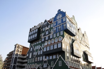 An architectural structure resembling a stack of traditional Dutch houses with varying facades and colors, featuring green, blue, and white hues often associated with classic Dutch architecture. The building's unique design gives the impression of multiple houses combined into a single unit.