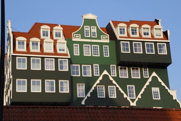 A unique building features traditional Dutch architectural styles with a modern twist. The facade has green walls, white-framed windows, and red-tiled roofs. Multiple gables and dormer windows add to the intricate design, creating a layered, stacked appearance.