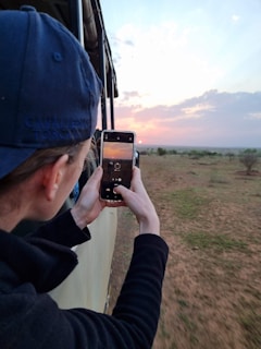 A person wearing a blue cap and black jacket is taking a photo of a sunset from an open vehicle. The landscape consists of a plain with sparse vegetation, and the sky is filled with pink and orange hues due to the sunset.
