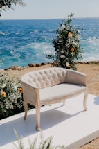 An elegant, vintage-style loveseat upholstered in light fabric is placed on a white platform. The loveseat is surrounded by floral arrangements with white and orange flowers. In the background, the deep blue ocean waves crash against a rocky shore, creating a serene coastal backdrop.