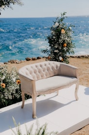 An elegant, vintage-style loveseat upholstered in light fabric is placed on a white platform. The loveseat is surrounded by floral arrangements with white and orange flowers. In the background, the deep blue ocean waves crash against a rocky shore, creating a serene coastal backdrop.