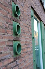 A brick wall with three green circular pipe-like structures protruding from it. The wall has green accents around a large window visible to the right.
