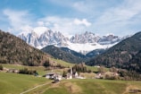 A picturesque landscape featuring a quaint Alpine village nestled in a lush, green valley surrounded by tall, evergreen forests. Snow-capped mountain peaks rise majestically in the background under a partly cloudy sky.