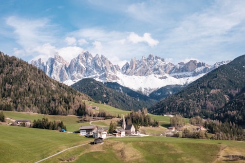 A picturesque landscape featuring a quaint Alpine village nestled in a lush, green valley surrounded by tall, evergreen forests. Snow-capped mountain peaks rise majestically in the background under a partly cloudy sky.