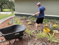 A person is engaged in gardening activities, using a rake to work on the flower bed in a landscaped yard. They are wearing headphones and casual clothing. Nearby, a black wheelbarrow labeled 'AMES' and a yellow bucket can be seen. The surrounding environment includes green plants, mulch, and a garden hose.