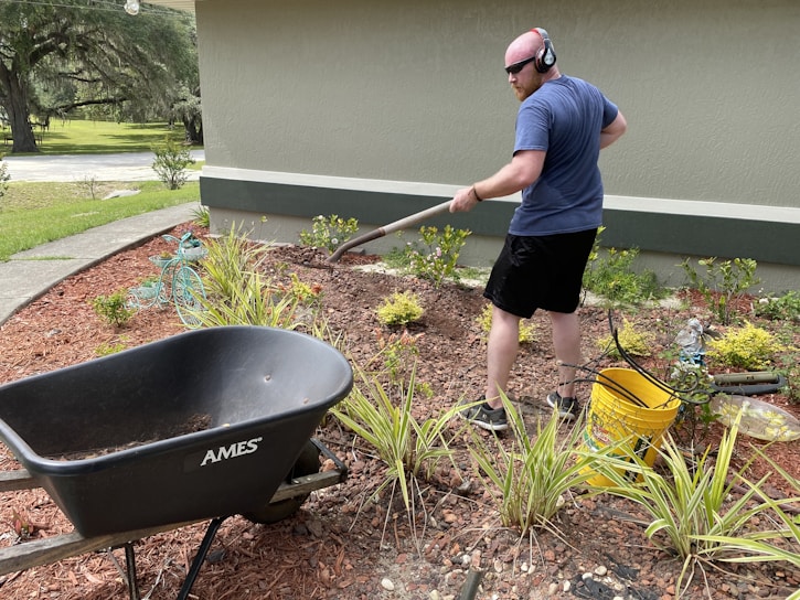 A person is engaged in gardening activities, using a rake to work on the flower bed in a landscaped yard. They are wearing headphones and casual clothing. Nearby, a black wheelbarrow labeled 'AMES' and a yellow bucket can be seen. The surrounding environment includes green plants, mulch, and a garden hose.