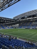 A modern sports stadium with a large, green soccer field and surrounding seating areas. Blue and yellow seats are visible, with some spectators scattered across the stands. The stadium is partially covered with a steel lattice roof, showing a clear blue sky. Several security personnel in high-visibility vests are present.
