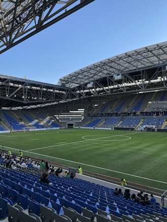 A modern sports stadium with a large, green soccer field and surrounding seating areas. Blue and yellow seats are visible, with some spectators scattered across the stands. The stadium is partially covered with a steel lattice roof, showing a clear blue sky. Several security personnel in high-visibility vests are present.