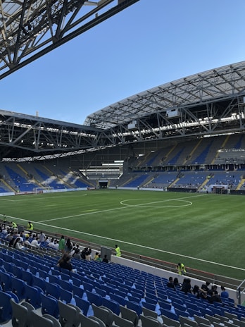 A modern sports stadium with a large, green soccer field and surrounding seating areas. Blue and yellow seats are visible, with some spectators scattered across the stands. The stadium is partially covered with a steel lattice roof, showing a clear blue sky. Several security personnel in high-visibility vests are present.