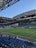 A modern sports stadium with a large, green soccer field and surrounding seating areas. Blue and yellow seats are visible, with some spectators scattered across the stands. The stadium is partially covered with a steel lattice roof, showing a clear blue sky. Several security personnel in high-visibility vests are present.