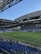 A modern sports stadium with a large, green soccer field and surrounding seating areas. Blue and yellow seats are visible, with some spectators scattered across the stands. The stadium is partially covered with a steel lattice roof, showing a clear blue sky. Several security personnel in high-visibility vests are present.