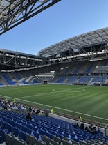 A modern sports stadium with a large, green soccer field and surrounding seating areas. Blue and yellow seats are visible, with some spectators scattered across the stands. The stadium is partially covered with a steel lattice roof, showing a clear blue sky. Several security personnel in high-visibility vests are present.