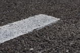 A close-up view of a rough-textured asphalt road with a white painted line running diagonally across it. The texture of the road surface is clearly visible, highlighting small stones and granules.