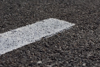 A close-up view of a rough-textured asphalt road with a white painted line running diagonally across it. The texture of the road surface is clearly visible, highlighting small stones and granules.