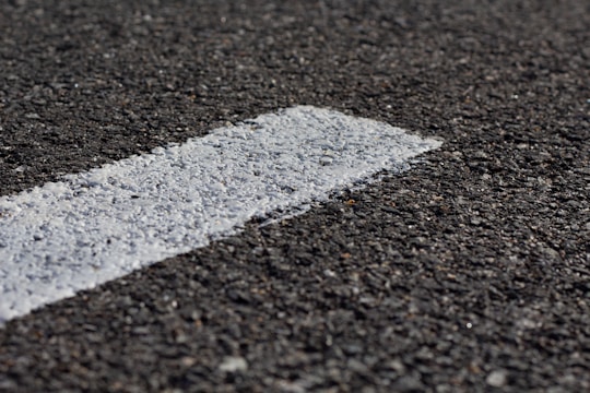 A close-up view of a rough-textured asphalt road with a white painted line running diagonally across it. The texture of the road surface is clearly visible, highlighting small stones and granules.