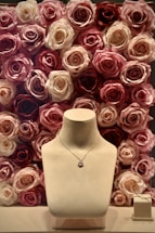 A jewelry display with a necklace on a stand, set against a background of roses in various shades of pink, cream, and red. To the side, a pair of delicate earrings is placed on a small box.