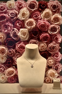 A jewelry display with a necklace on a stand, set against a background of roses in various shades of pink, cream, and red. To the side, a pair of delicate earrings is placed on a small box.