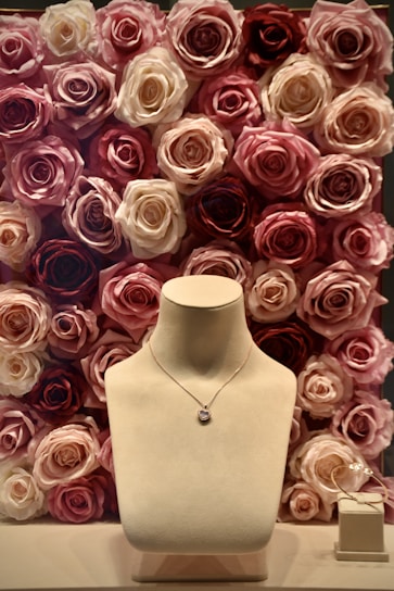 A jewelry display with a necklace on a stand, set against a background of roses in various shades of pink, cream, and red. To the side, a pair of delicate earrings is placed on a small box.