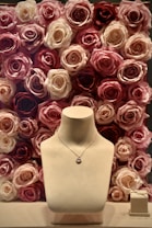 A jewelry display with a necklace on a stand, set against a background of roses in various shades of pink, cream, and red. To the side, a pair of delicate earrings is placed on a small box.