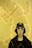 A mannequin dressed in stylish clothing is set against a bright yellow background with a large, abstract soft sculpture. The mannequin is wearing a black bucket hat with a logo, white sunglasses, a zip-up outfit, and a dark jacket.