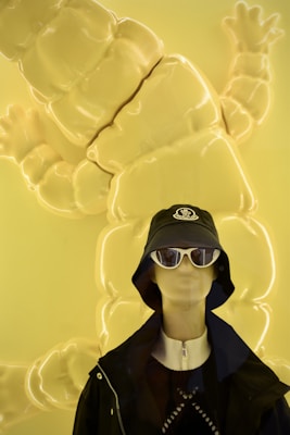 A mannequin dressed in stylish clothing is set against a bright yellow background with a large, abstract soft sculpture. The mannequin is wearing a black bucket hat with a logo, white sunglasses, a zip-up outfit, and a dark jacket.