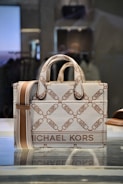 A stylish designer handbag made from a textured material featuring a repeating geometric pattern with the brand name prominently displayed. The colors consist of subtle shades of brown and beige, creating a sophisticated look. The bag is placed on a reflective surface in a well-lit setting.
