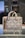 A stylish designer handbag made from a textured material featuring a repeating geometric pattern with the brand name prominently displayed. The colors consist of subtle shades of brown and beige, creating a sophisticated look. The bag is placed on a reflective surface in a well-lit setting.