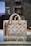 A stylish designer handbag made from a textured material featuring a repeating geometric pattern with the brand name prominently displayed. The colors consist of subtle shades of brown and beige, creating a sophisticated look. The bag is placed on a reflective surface in a well-lit setting.