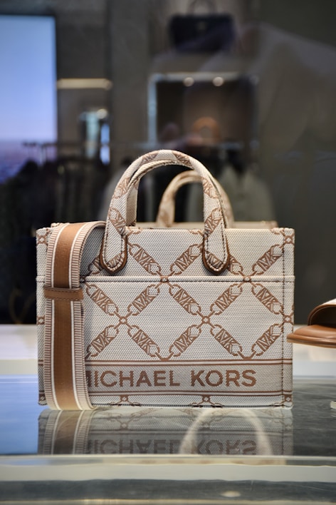 A stylish designer handbag made from a textured material featuring a repeating geometric pattern with the brand name prominently displayed. The colors consist of subtle shades of brown and beige, creating a sophisticated look. The bag is placed on a reflective surface in a well-lit setting.