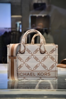 A stylish designer handbag made from a textured material featuring a repeating geometric pattern with the brand name prominently displayed. The colors consist of subtle shades of brown and beige, creating a sophisticated look. The bag is placed on a reflective surface in a well-lit setting.