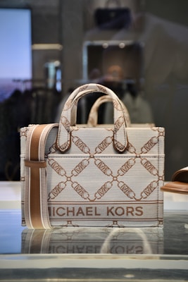 A stylish designer handbag made from a textured material featuring a repeating geometric pattern with the brand name prominently displayed. The colors consist of subtle shades of brown and beige, creating a sophisticated look. The bag is placed on a reflective surface in a well-lit setting.