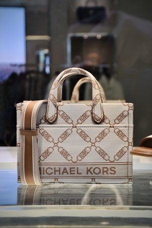 A stylish designer handbag made from a textured material featuring a repeating geometric pattern with the brand name prominently displayed. The colors consist of subtle shades of brown and beige, creating a sophisticated look. The bag is placed on a reflective surface in a well-lit setting.