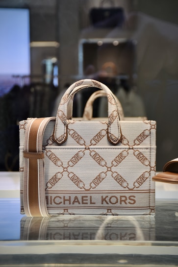 A stylish designer handbag made from a textured material featuring a repeating geometric pattern with the brand name prominently displayed. The colors consist of subtle shades of brown and beige, creating a sophisticated look. The bag is placed on a reflective surface in a well-lit setting.
