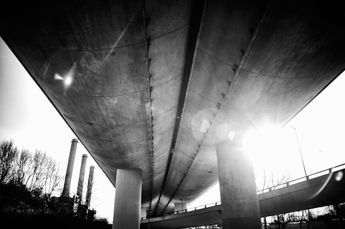 A black and white photograph captured from beneath a large bridge or overpass, showing the structural details and beams. The sunlight is visible in the background creating lens flare effects, and there are silhouettes of trees and industrial chimneys on one side.